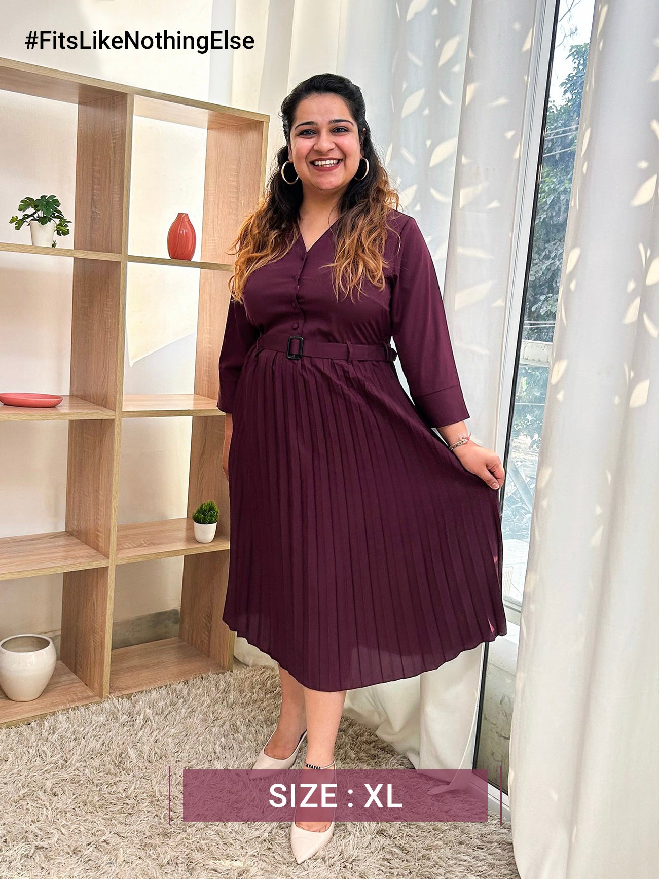 A woman standing in a room wearing a maroon A-line pleated dress with a shirt collar, three-quarter sleeves, and a belt.