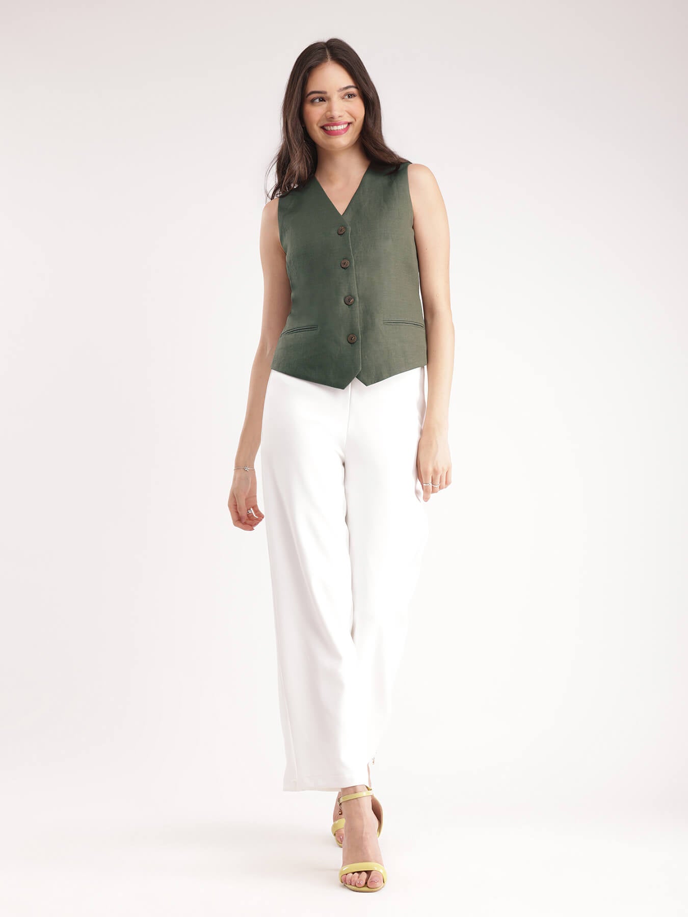 A woman wearing a green linen waistcoat with V-neck and 2 front pockets, paired with white trousers.