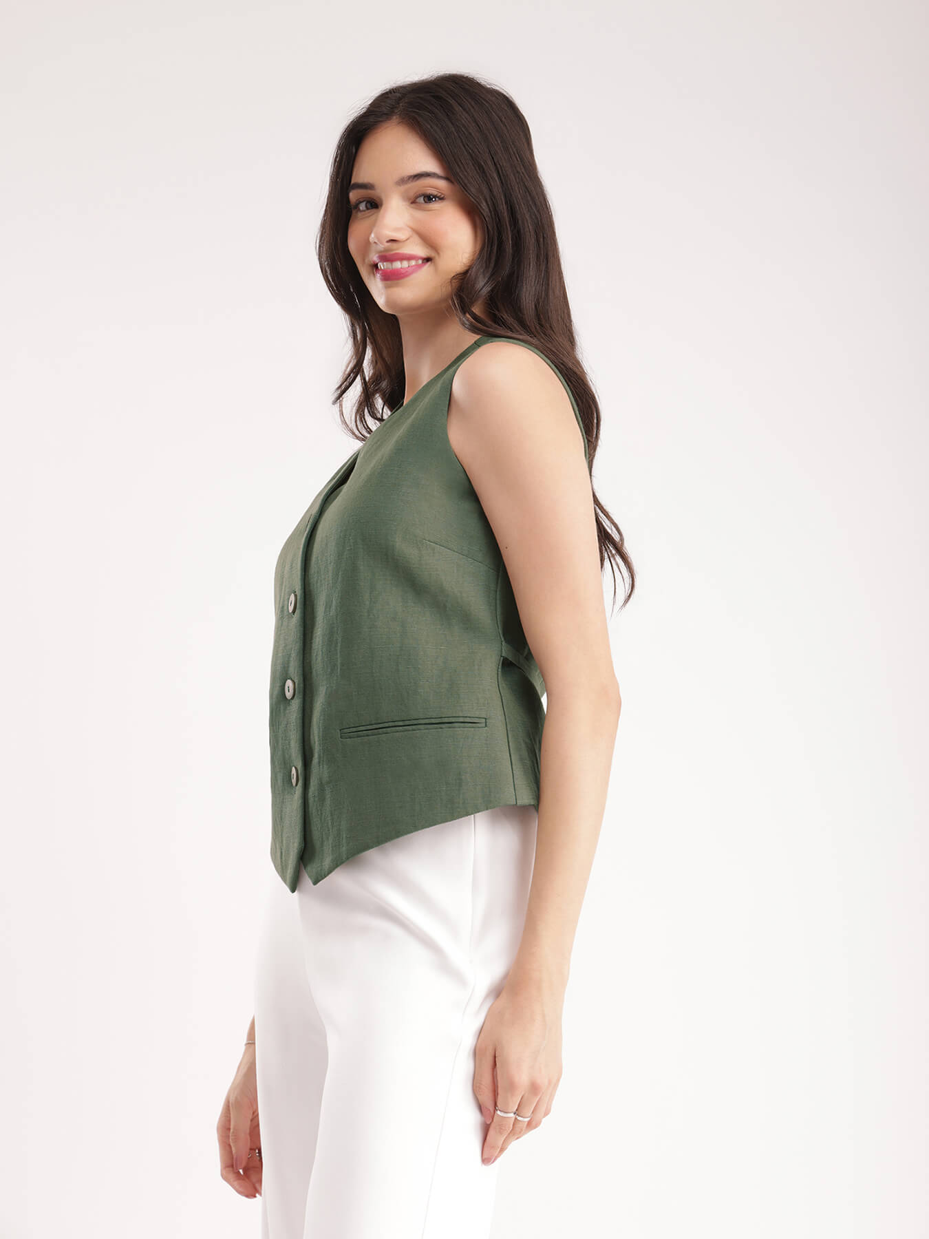 A woman wearing a green linen waistcoat with V-neck and 2 front pockets, paired with white trousers.