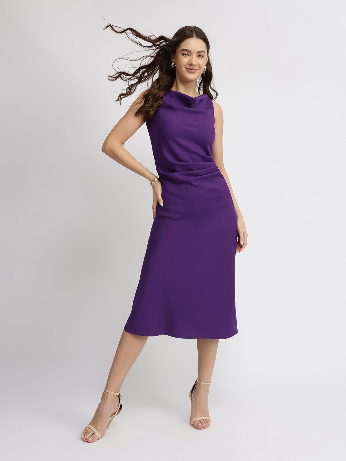 A woman modeling a purple pleated midi dress with a cowl neck and A-line silhouette.