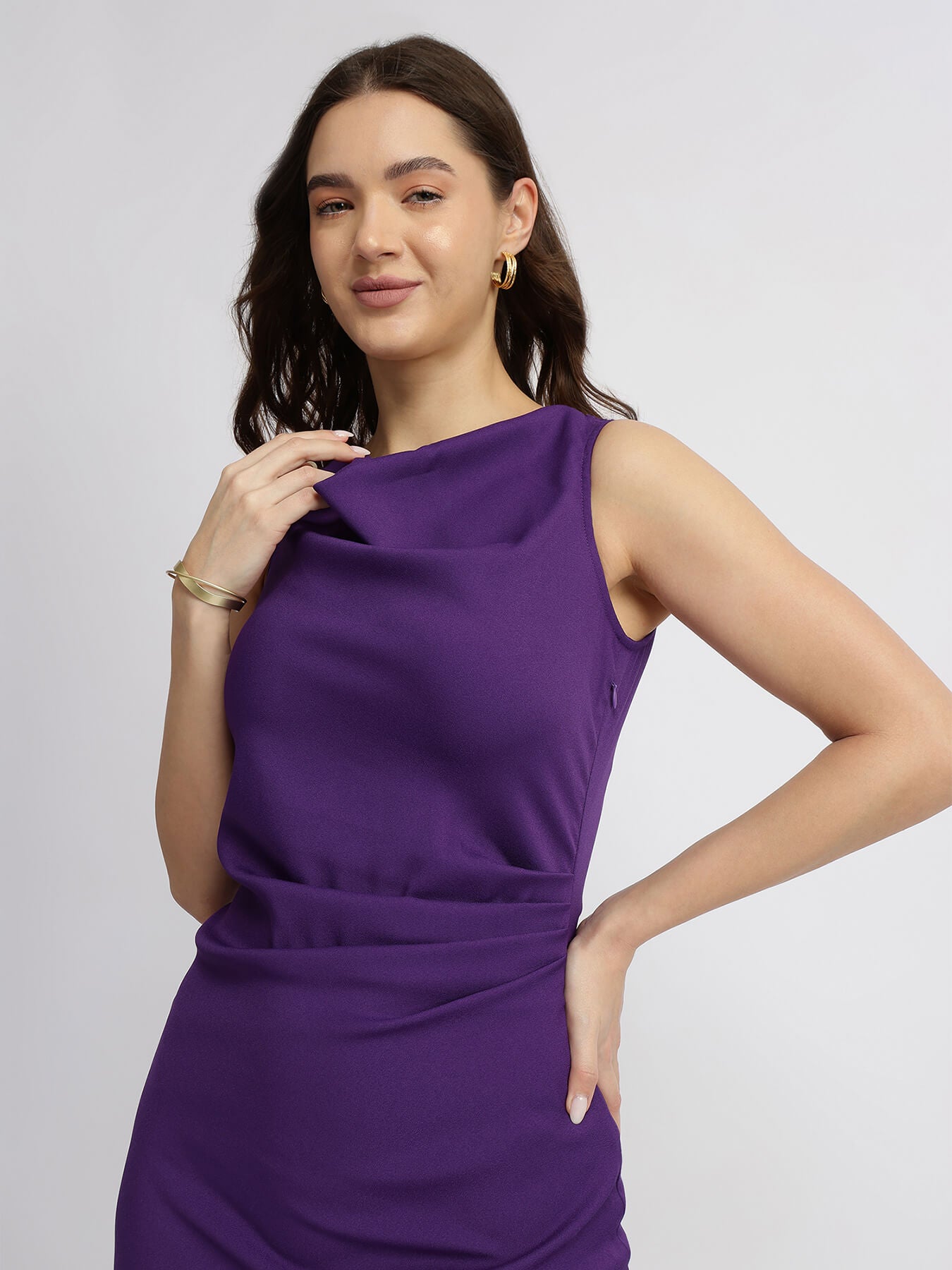 A woman wearing a purple pleated midi dress with a boat neckline, sleeveless design, and falling pleats at the skirt.