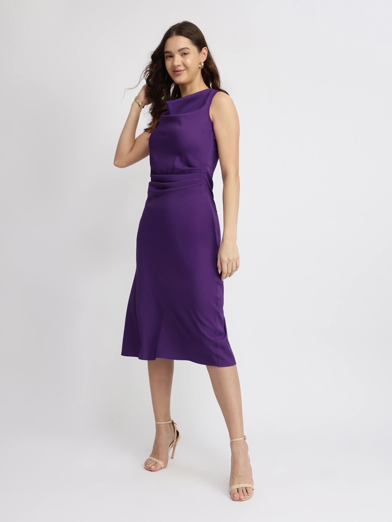 A woman wearing a purple pleated midi dress with a boat neckline, sleeveless design, and falling pleats at the skirt.
