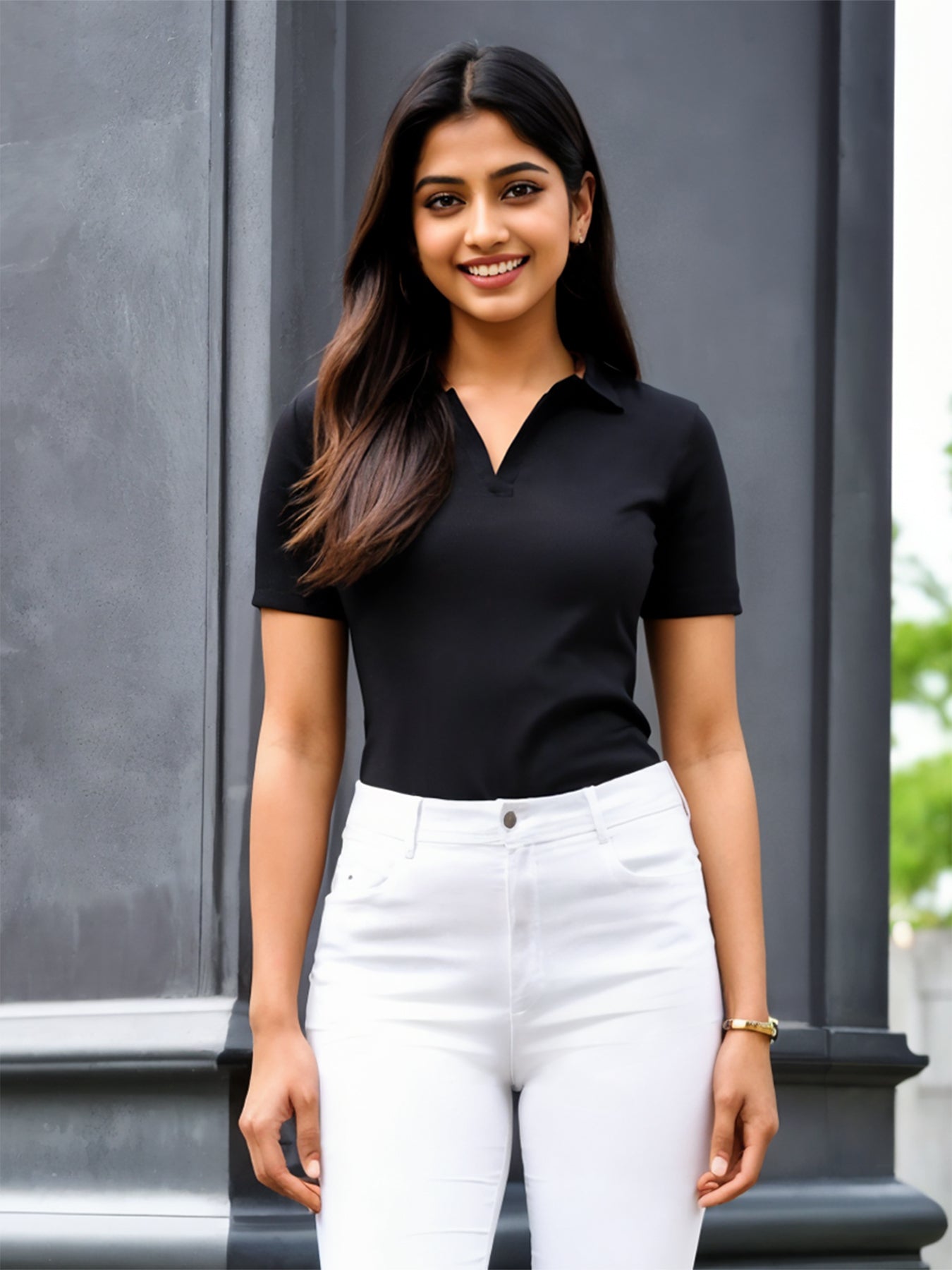 A woman wearing a black collared t-shirt with short sleeves and white pants standing against a gray background.