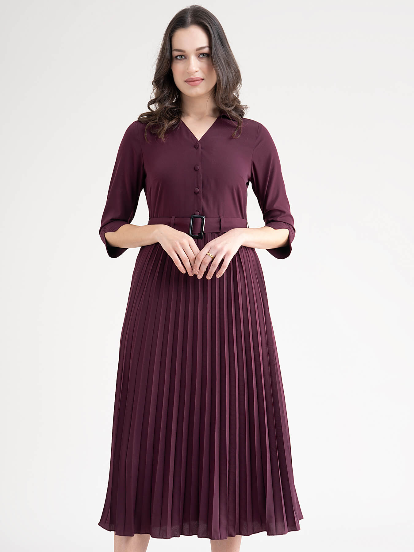 A woman standing in a room wearing a maroon A-line pleated dress with a shirt collar, three-quarter sleeves, and a belt.