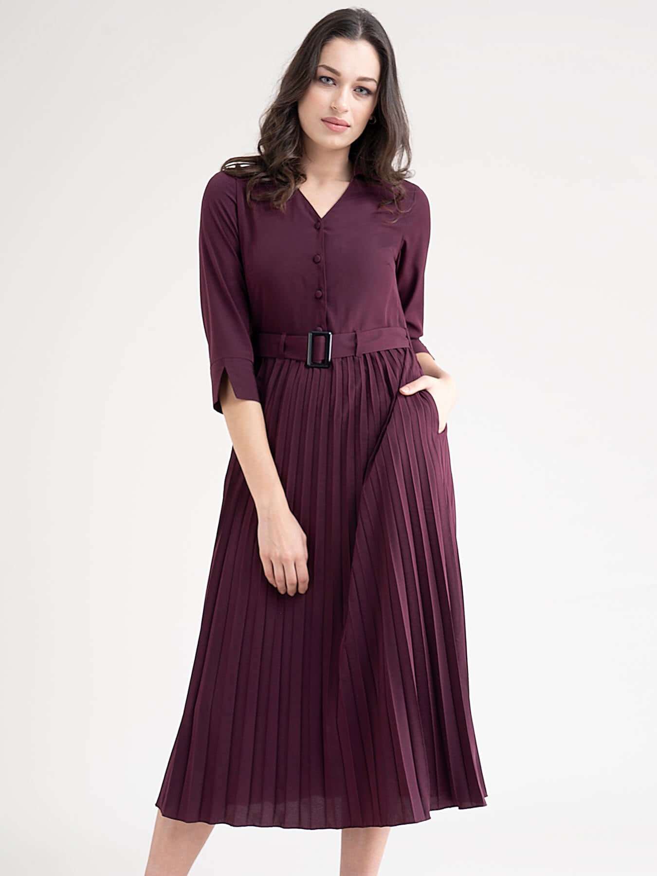 A woman standing in a room wearing a maroon A-line pleated dress with a shirt collar, three-quarter sleeves, and a belt.