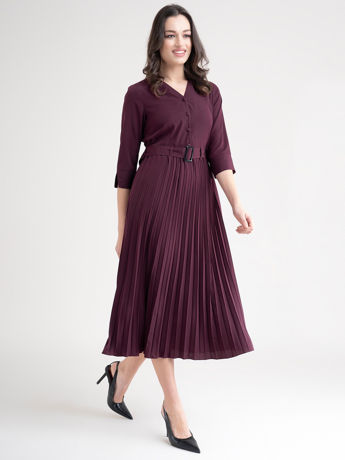 A woman standing in a room wearing a maroon A-line pleated dress with a shirt collar, three-quarter sleeves, and a belt.