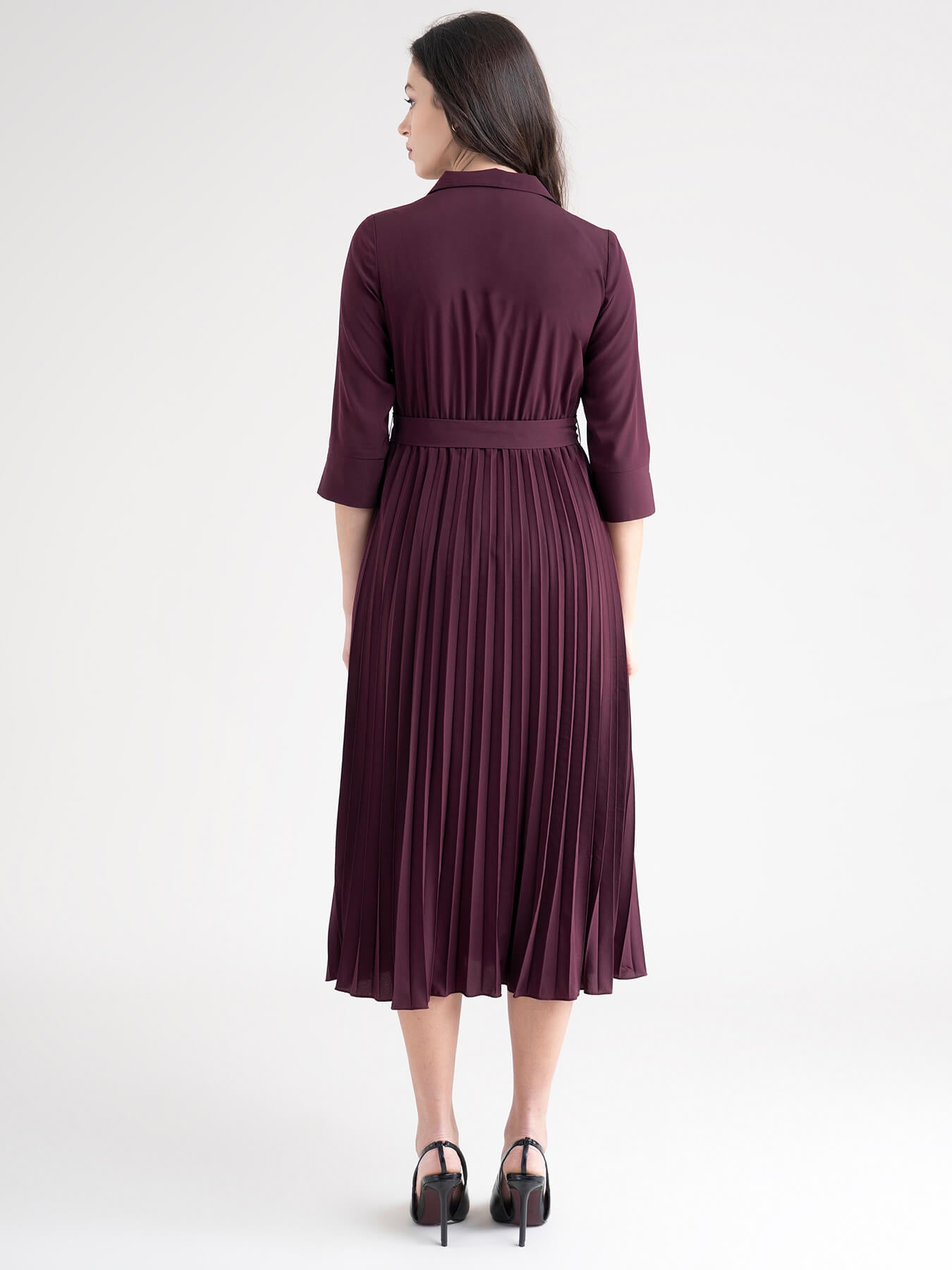 A woman standing in a room wearing a maroon A-line pleated dress with a shirt collar, three-quarter sleeves, and a belt.