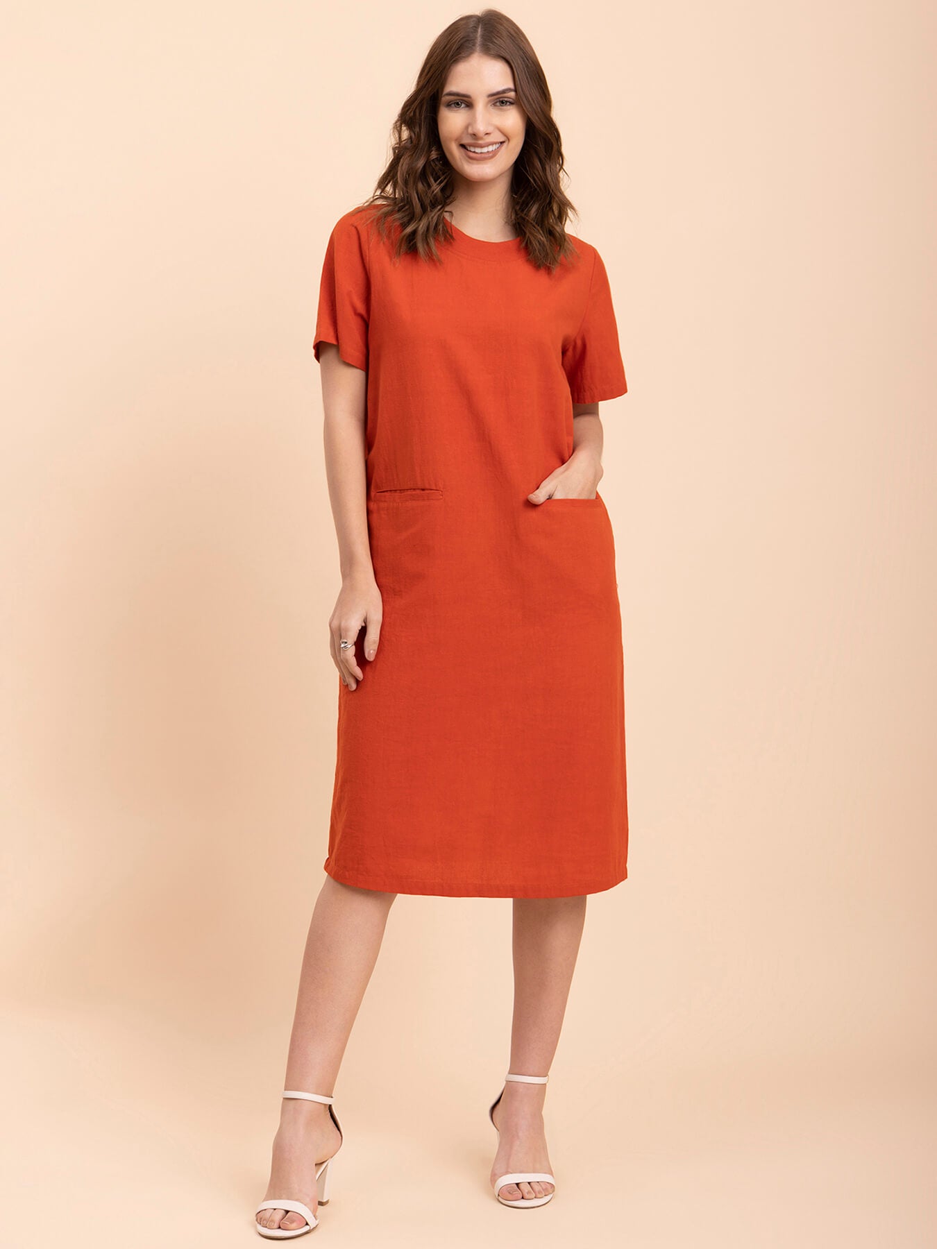 A woman standing in a burnt orange shift dress with short sleeves and a round neckline, paired with white ankle strap sandals.