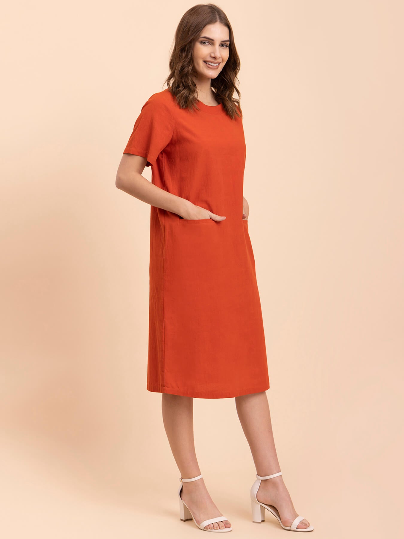 A woman standing in a burnt orange shift dress with short sleeves and a round neckline, paired with white ankle strap sandals.