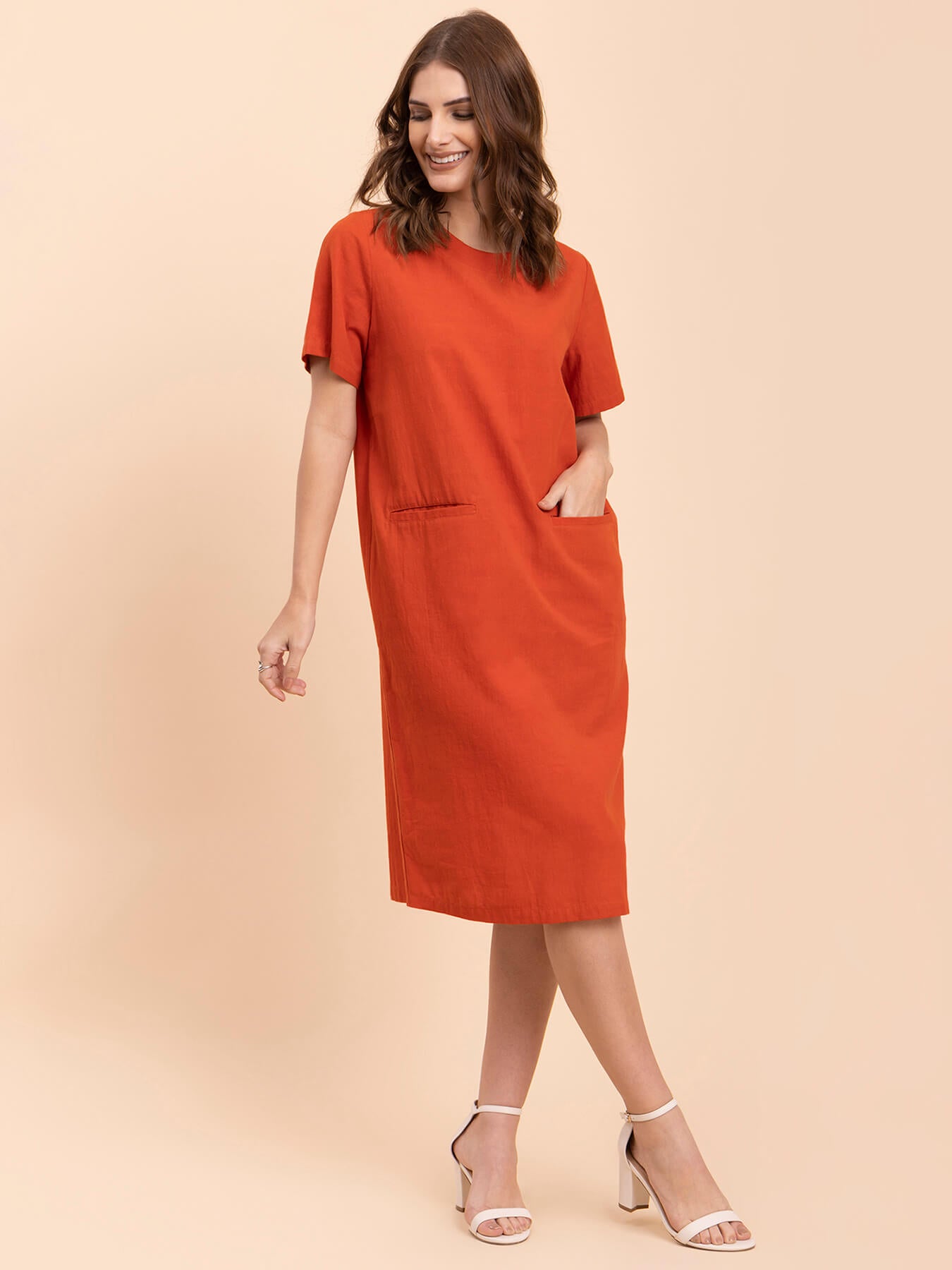 A woman standing in a burnt orange shift dress with short sleeves and a round neckline, paired with white ankle strap sandals.