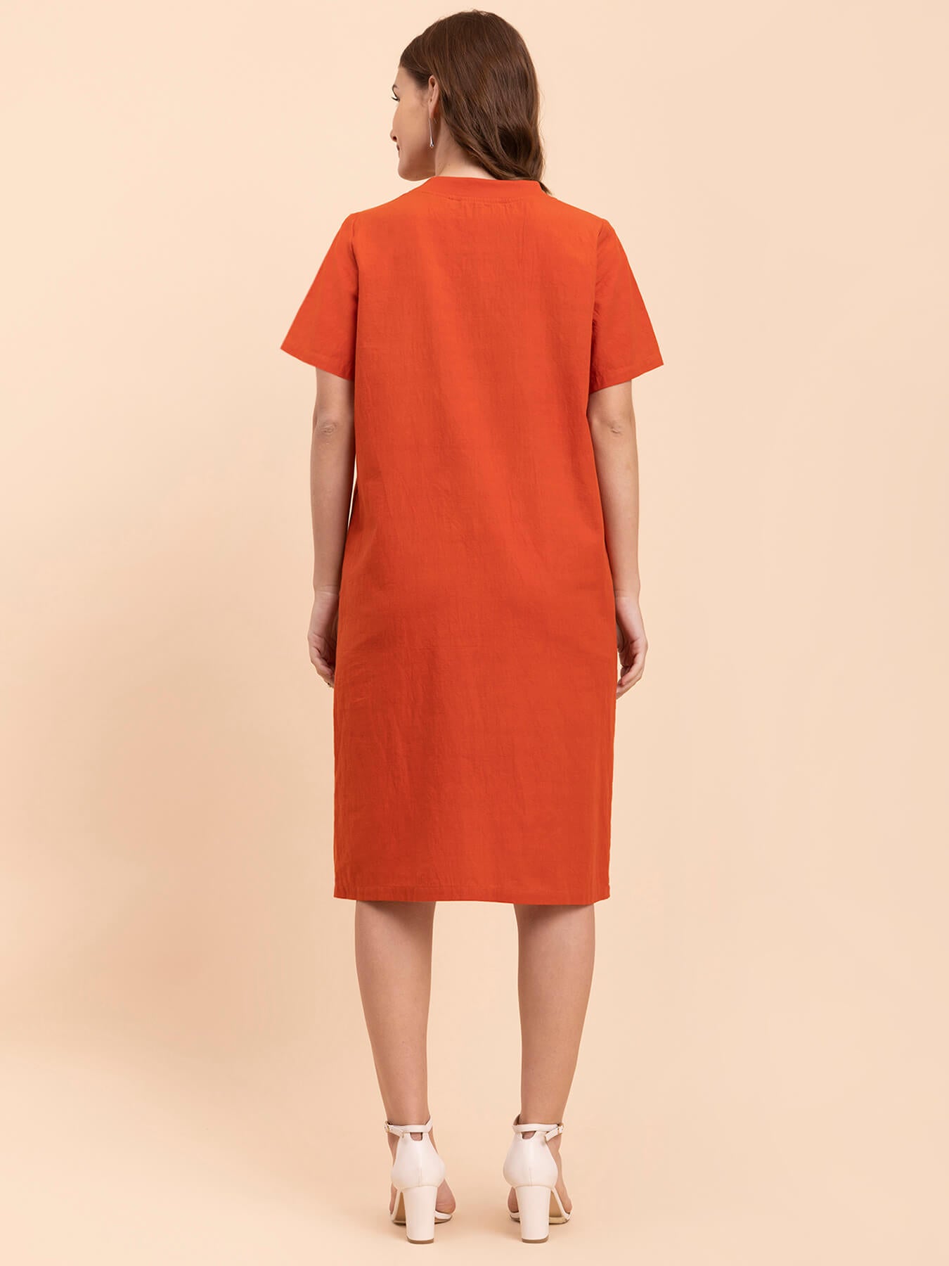A woman standing in a burnt orange shift dress with short sleeves and a round neckline, paired with white ankle strap sandals.
