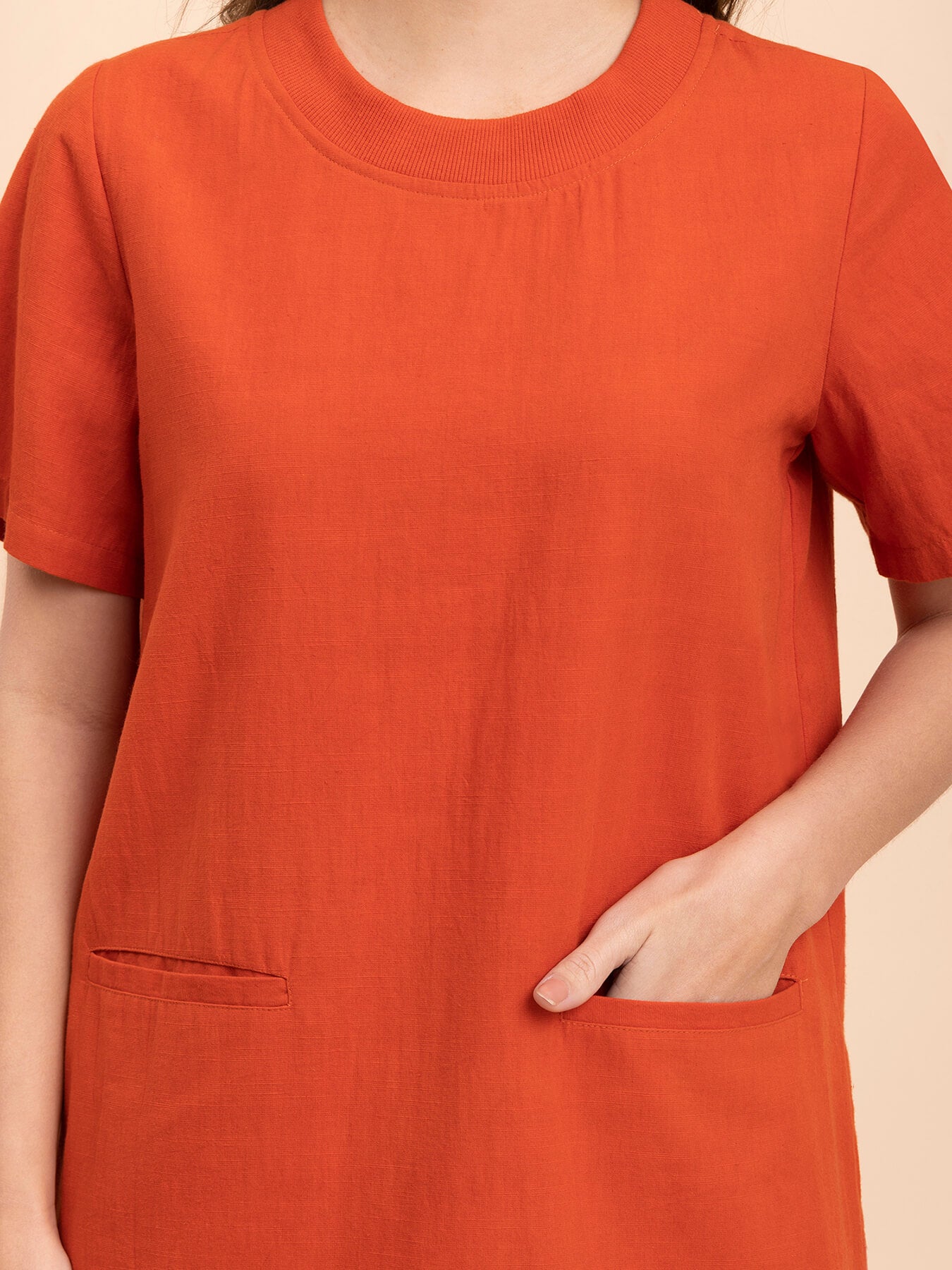 A woman standing in a burnt orange shift dress with short sleeves and a round neckline, paired with white ankle strap sandals.